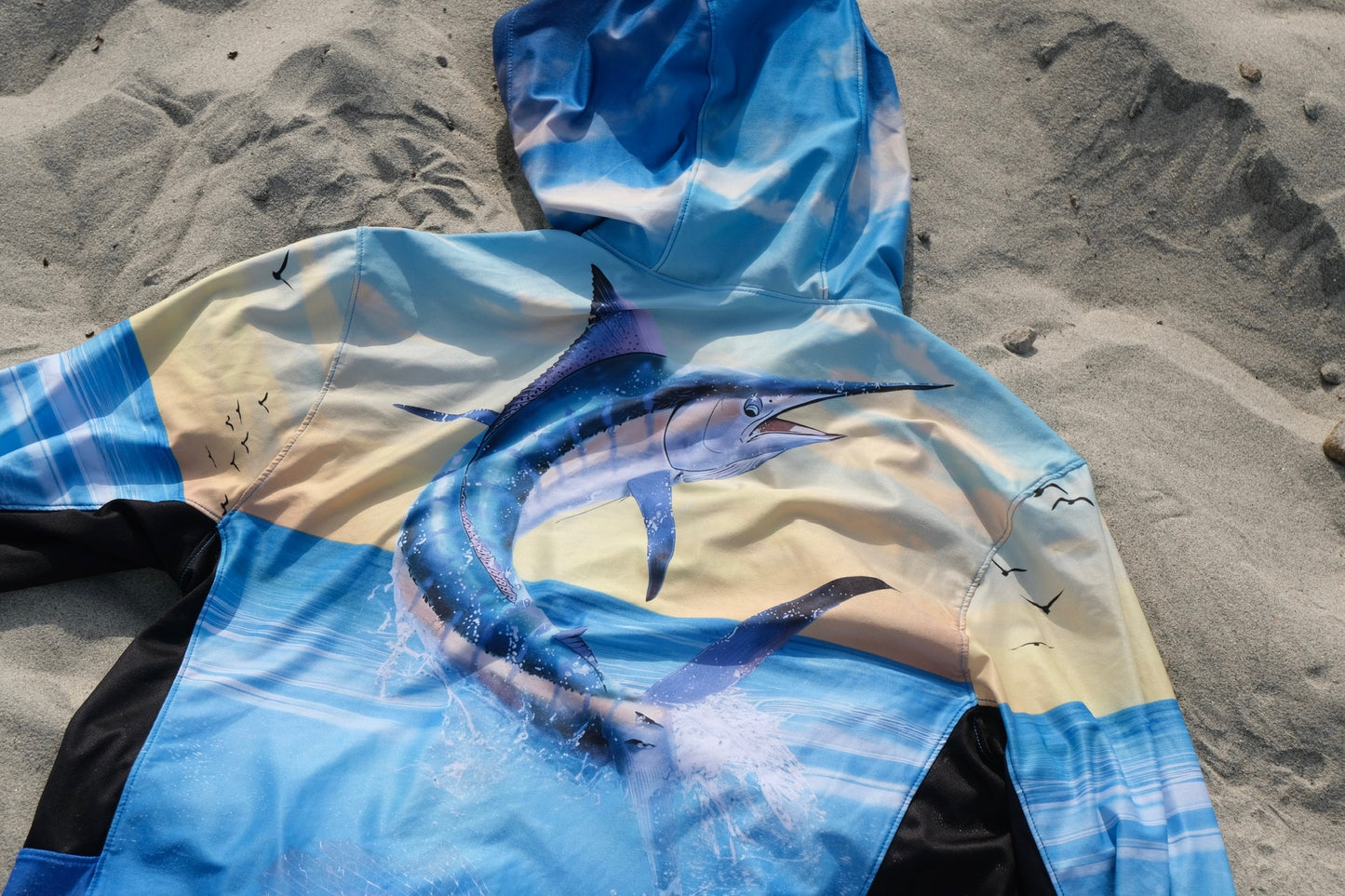 Angler 24-7 hoodie with marlin fish design on sandy beach.
